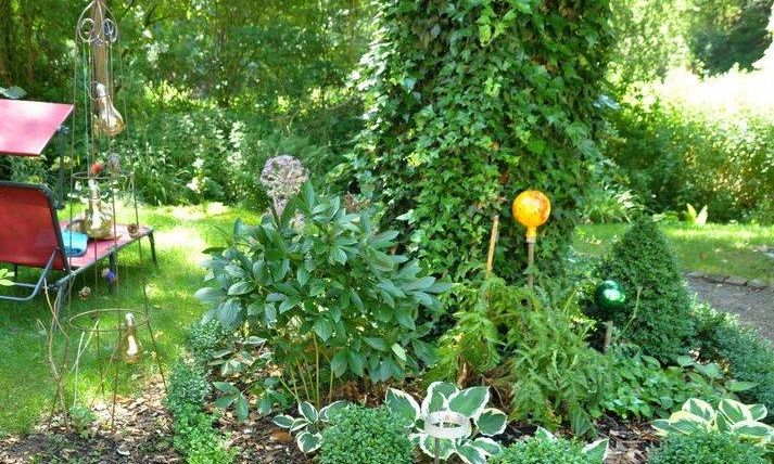 Ein schöner Garten mit grünen Pflanzen und einem begrünten Baum. Im Hintergrund sieht man eine Liege und dekorative Gartenleuchten.