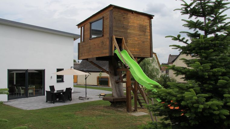 Une maison de jeu en bois avec un toboggan vert se trouve dans le jardin. À côté, il y a un espace de vie moderne et une terrasse.