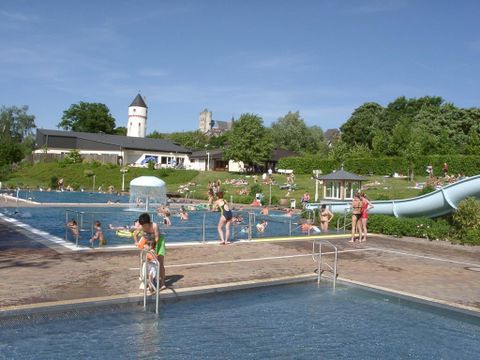 Ein Schwimmbad mit mehreren Becken und Wasserrutschen. Viele Menschen genießen den sonnigen Tag am Wasser.