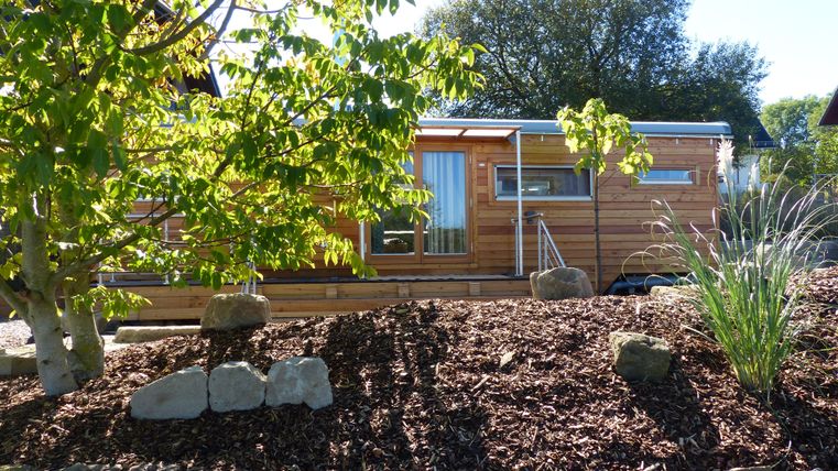 Ein modernes Holzhaus umgeben von Bäumen und Sträuchern. Der Garten ist gepflegt mit Kies und Steinmauern.