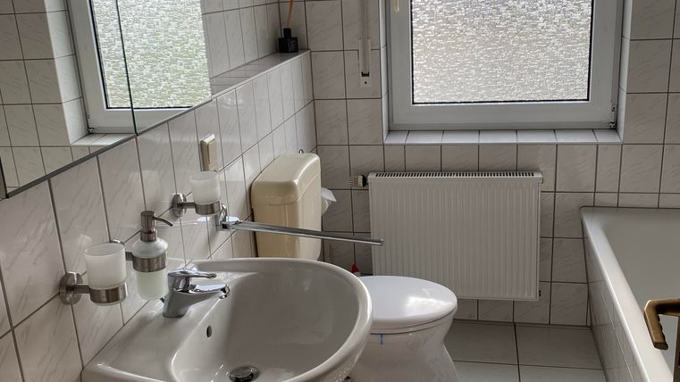 Ein modernes Badezimmer mit weißen Fliesen und einem großen Spiegel. Es gibt ein Waschbecken, eine Toilette und eine Badewanne, sowie ein Fenster mit Sichtschutz.
