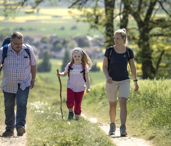 Ein Mann, eine Frau und ein Kind wandern auf einem l&auml;ndlichen Weg. Das Kind tr&auml;gt einen Wanderstock. Im Hintergrund sind Felder und B&auml;ume zu sehen., &copy; Kappest, REMET