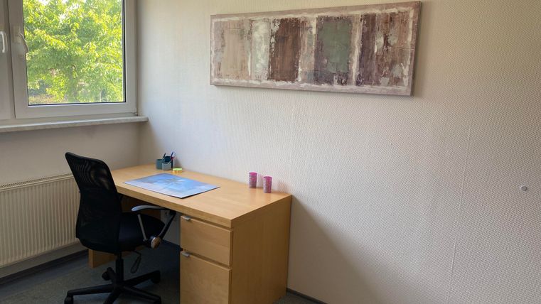 Ein einfaches Büro mit einem Schreibtisch und einem Stuhl. An der Wand hängt ein Gemälde, und durch das Fenster ist Tageslicht sichtbar.