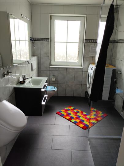 Ein modernes Badezimmer mit einer Dusche und einem Waschbecken. Auf dem Boden liegt ein bunter Teppich aus rechteckigen Mustern.