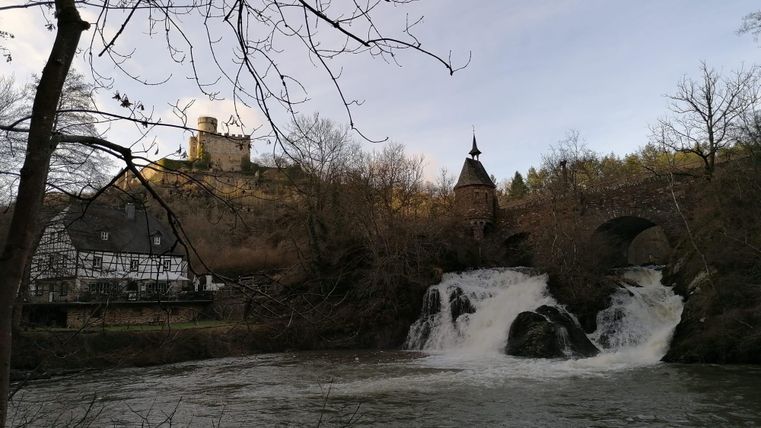Ein malerischer Wasserfall fließt vor einer alten Burg. Im Hintergrund sind Bäume und ein historisches Gebäude zu sehen.