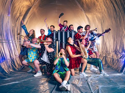 Eine Gruppe von Musikern posiert in bunten Kostümen in einem glitzernden Raum. Sie halten verschiedene Musikinstrumente und strahlen Freude aus.