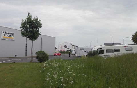 Eine Ansicht von einem Parkplatz mit einem Wohnmobil und grünem Gras. Im Hintergrund sind einige Gebäude und ein Schild von Niessmann + Bischoff zu sehen.
