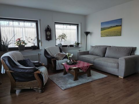 Ein gemütliches Wohnzimmer mit hellen Fenstern und Pflanzen. Es gibt eine graue Couch und bequeme Stühle, die um einen Tisch angeordnet sind.