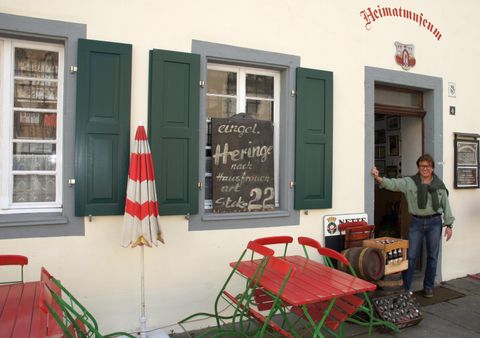 Een restaurant met groene luiken en roodachtige tafels buiten. Iemand staat voor de deur en verwelkomt de gasten.