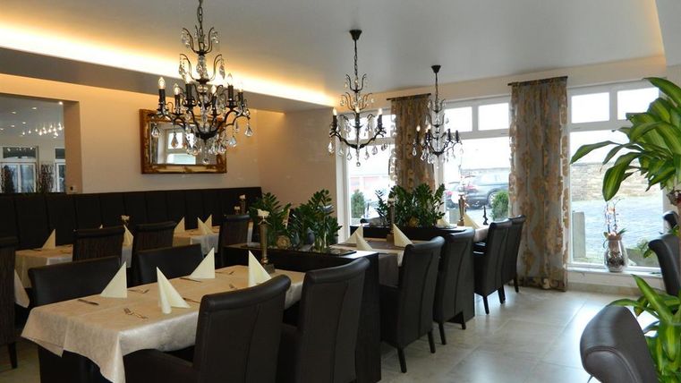 Ein elegantes Restaurant mit dunklen Holzmöbeln und stilvollen Kronleuchtern. Pflanzen und dekorative Vorhänge schaffen eine gemütliche Atmosphäre.