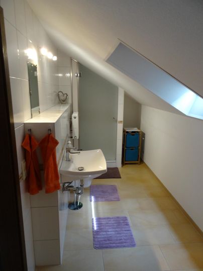 Ein modernes Badezimmer mit schrägen Wänden und einem großen Fenster. Es gibt einen Waschbecken, Handtücher und dekorative Badmatten.