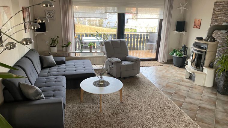 Ein gemütliches Wohnzimmer mit einem grauen Sofa und einem Relaxsessel. Der Raum hat große Fenster und einen modernen Kamin.