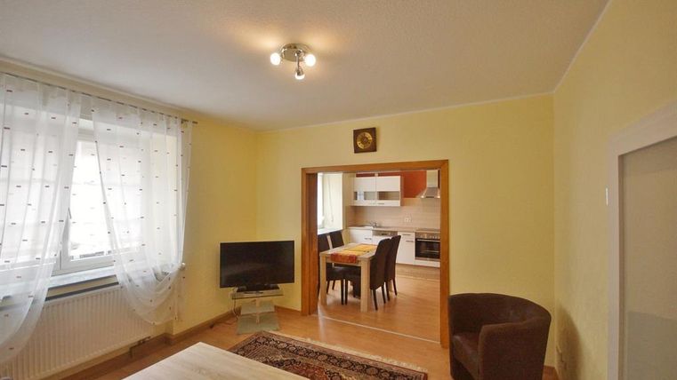 Ein gemütliches Wohnzimmer mit einem Fernseher, einem Tisch und einem Sessel. Im Hintergrund befindet sich ein Essbereich mit einem Tisch und Stühlen.