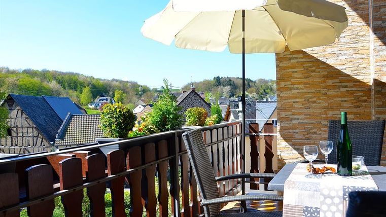 Ein gemütlicher Balkon mit einem Sonnenschirm, einem Tisch und Stühlen. Im Hintergrund sind grüne Hügel und traditionelle Häuser zu sehen.