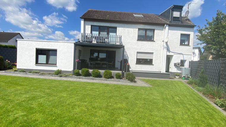 Ein modernes, zweigeschossiges Haus mit einer gepflegten Rasenfläche und bunten Blumen. Der Himmel ist blau mit einigen Wolken.
