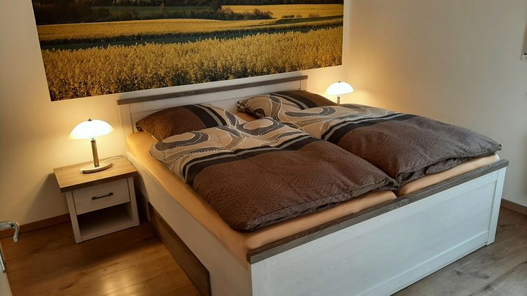 Une chambre confortable avec un lit double et des lumières douces. En arrière-plan, il y a un grand tableau de paysage avec des champs.