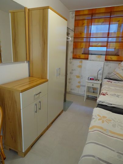 Een eenvoudige kamer met twee bedden en een beige kast. De ramen zijn versierd met oranje gordijnen.