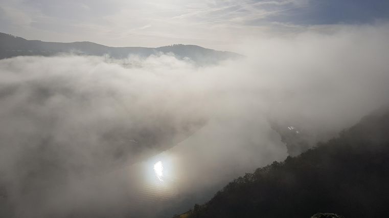 Eine neblige Landschaft mit einem ruhigen Fluss, der das Licht der Sonne reflektiert. Im Hintergrund sind sanfte Hügel und ein blauer Himmel zu sehen.