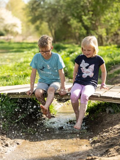 Deux enfants sont assis sur un pont en bois et barbotent avec leurs pieds dans l'eau. En arrière-plan, on voit des prairies vertes et des arbres.
