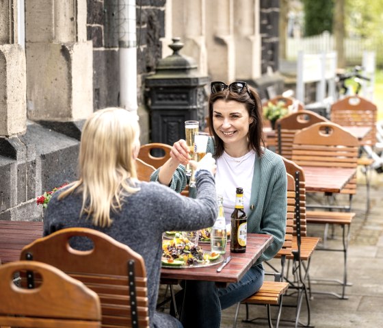 Zwei Frauen sitzen an einem Holztisch im Freien und sto&szlig;en mit Getr&auml;nken an. Im Hintergrund sind Fahrr&auml;der zu sehen., &copy; Eifel Tourismus GmbH, Dominik Ketz