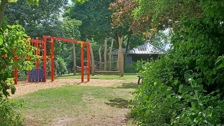 Ein Spielplatz mit roten Spielgeräten und einem Kletterbaum. Umgeben von viel Grün und Bäumen.