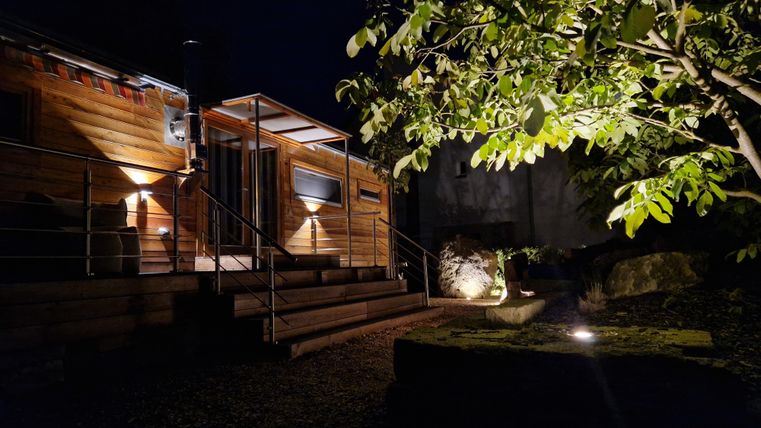 Ein hübsches Holzhaus bei Nacht, sanft beleuchtet von verschiedenen Lichtern. Umgeben von einem gepflegten Garten mit einem Baum im Vordergrund.