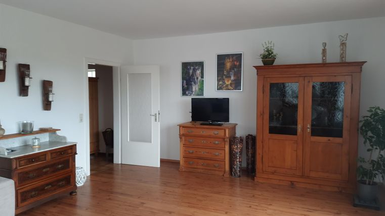 Ein helles Wohnzimmer mit holzgetäfeltem Boden und modernen Möbeln. An der Wand hängen Bilder und es gibt eine Pflanze in der Ecke.