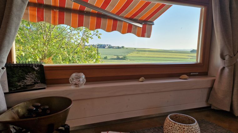 Ein gemütlicher Raum mit Blick auf die grüne Landschaft. Die Fenster mit rot-weiß gestreiften Markisen lassen viel Licht herein.