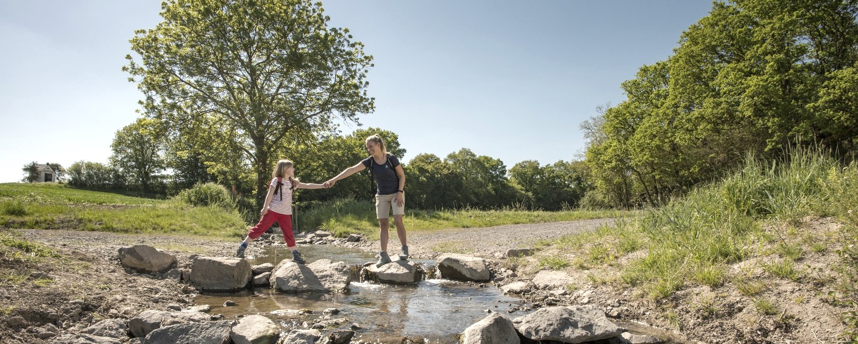 Zwei Personen &uuml;berqueren einen kleinen Bach auf Steinen. Im Hintergrund sind B&auml;ume und ein klarer Himmel zu sehen., &copy; Kappest, REMET