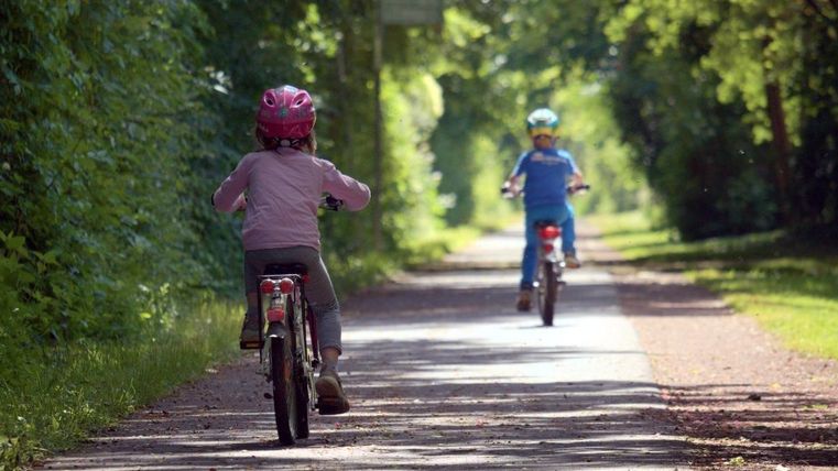Zwei Kinder fahren auf Fahrrädern einen schmalen Weg entlang. Umgeben von Bäumen genießen sie das schöne Wetter.