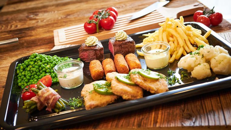 Ein appetitliches Gericht auf einem Tablett mit Rindersteaks, Kroketten, Pommes frites und verschiedenen Gemüsebeilagen. Dazu gibt es zwei Saucen in kleinen Schalen.
