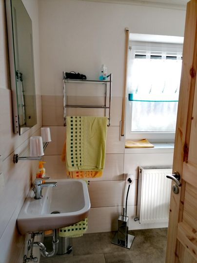 Ein modernes Badezimmer mit einem Waschbecken und einem Handtuchhalter. Die Fenster lassen viel Tageslicht herein und die Farben sind in sanften Tönen gehalten.