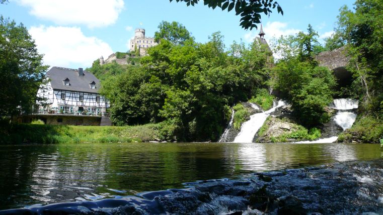 Ein malerischer Wasserfall fließt in einen ruhigen Fluss. Im Hintergrund sind Bäume und ein Schloss zu sehen, unter einem klaren blauen Himmel.