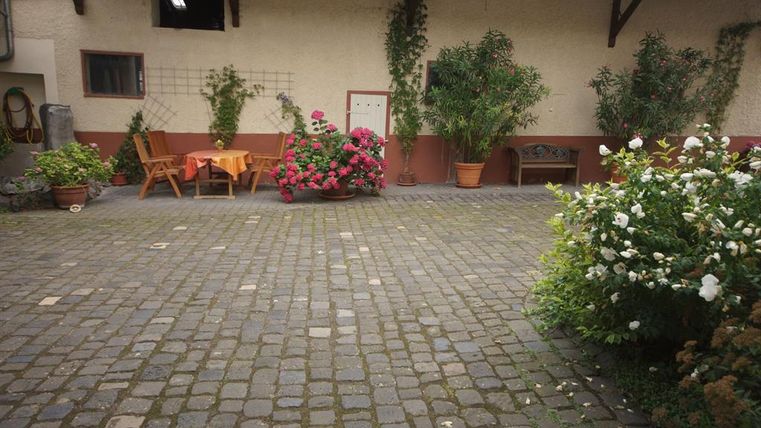 Ein gemütlicher Innenhof mit Steinpflaster. Es gibt Pflanzen und eine Sitzgelegenheit aus Holz, umgeben von bunten Blumen.