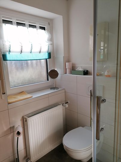 Ein helles Badezimmer mit einem Fenster, einem WC und einer Dusche. Auf dem Fensterbrett stehen Handtücher und Pflegeprodukte.