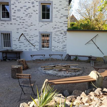 Ein gemütlicher Innenhof mit einem Feuerplatz und umgeben von Bänken. Die Wände sind schlicht und es gibt einige Werkzeuge zur Dekoration.