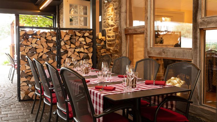 Ein einladender Außenbereich eines Restaurants mit einem Tisch, der für mehrere Gäste gedeckt ist. Im Hintergrund sind Holzscheite dekorativ gestapelt.