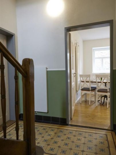 Un couloir lumineux avec un escalier et une porte ouverte sur une salle à manger. Le sol est carrelé avec un motif décoratif.