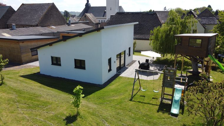 Une maison moderne et blanche avec un jardin attenant. À l'arrière-plan, on peut voir des équipements de jeu tels qu'une balançoire et un toboggan.