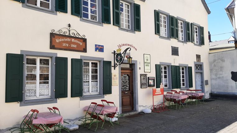 Ein traditionelles Restaurant mit einem einladenden Eingangsbereich und grünen Fensterläden. Draußen stehen rote Stühle und Tische, bereit für Gäste.