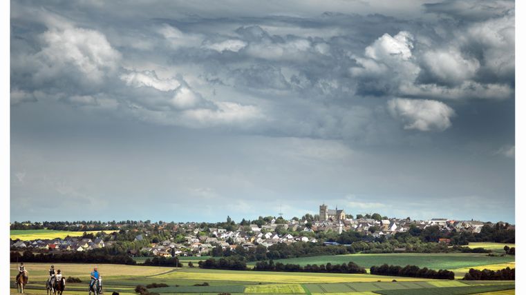 Eine malerische Landschaft mit sanften Hügeln und einer kleinen Stadt im Hintergrund. Der Himmel ist bewölkt und es sind einige Reiter in der Nähe zu sehen.
