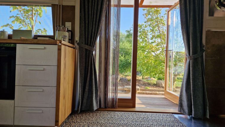 Eine helle Wohnküche mit einem offenen Eingang zu einem grünen Garten. Helle Vorhänge lassen Licht herein und schaffen eine einladende Atmosphäre.