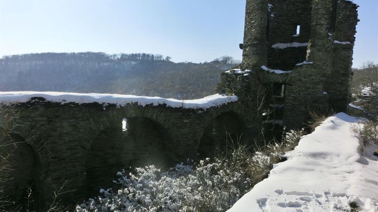 Une vieille ruine entourée de neige avec de petites fenêtres en arc. En arrière-plan, on aperçoit des collines boisées.