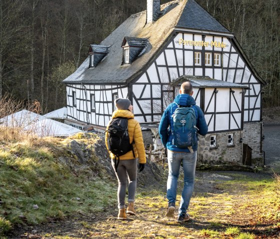 Zwei Personen wandern auf einem Pfad zur Pyrmonter Mühle. Die Fachwerkarchitektur der Mühle ist im Hintergrund sichtbar, umgeben von Wald., © Eifel Tourismus GmbH, D. Ketz
