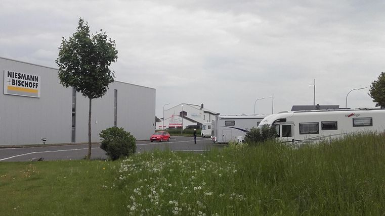 Eine Ansicht von einem Parkplatz mit einem Wohnmobil und grünem Gras. Im Hintergrund sind einige Gebäude und ein Schild von Niessmann + Bischoff zu sehen.