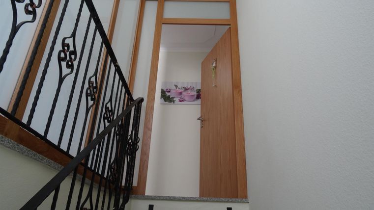 Eine Treppe mit einem schmiedeeisernen Geländer führt zu einer Holztür. Hinter der Tür ist ein Zimmer mit einem Bild an der Wand sichtbar.
