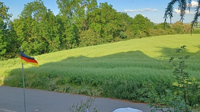 Ein ruhiger Außensitzbereich mit Tischen und Stühlen. Im Hintergrund erstrecken sich grüne Felder und Bäume unter einem blauen Himmel.