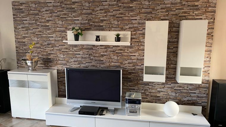 Ein moderner Wohnbereich mit einem weißen Fernsehmöbel und dekorativen Regalen. Die Wand ist mit einem ansprechenden Steinmuster verkleidet, und eine Pflanze sorgt für einen grünen Akzent.