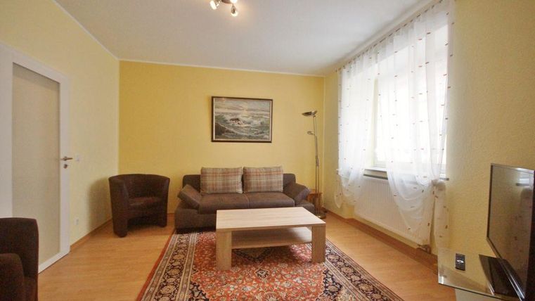 Ein gemütliches Wohnzimmer mit beiger Wandfarbe. Es gibt ein Sofa, zwei Sessel, einen Couchtisch und einen Teppich.