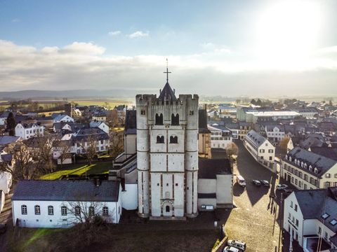 Ein historisches Gebäude mit einem hohen Turm in einer kleinen Stadt. Im Hintergrund sind weitere Häuser und eine ländliche Landschaft zu sehen.